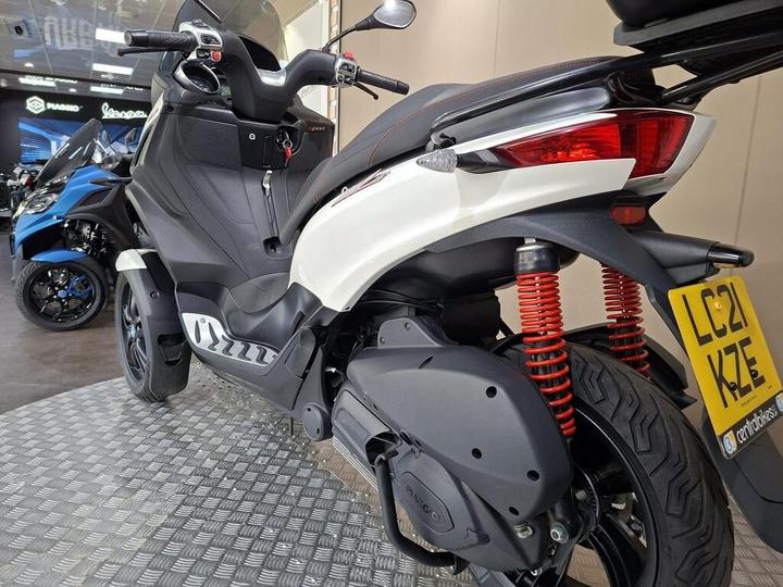 A white and black scooter with red shock absorbers is parked on a textured floor in a showroom, featuring a visible license plate reading "LC21 KZE." Other scooters are blurred in the background.