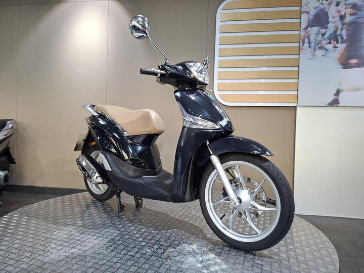 A sleek black scooter with a tan seat, showcased on a textured display surface, with a modern backdrop featuring a wooden design and a blurred image of people walking.