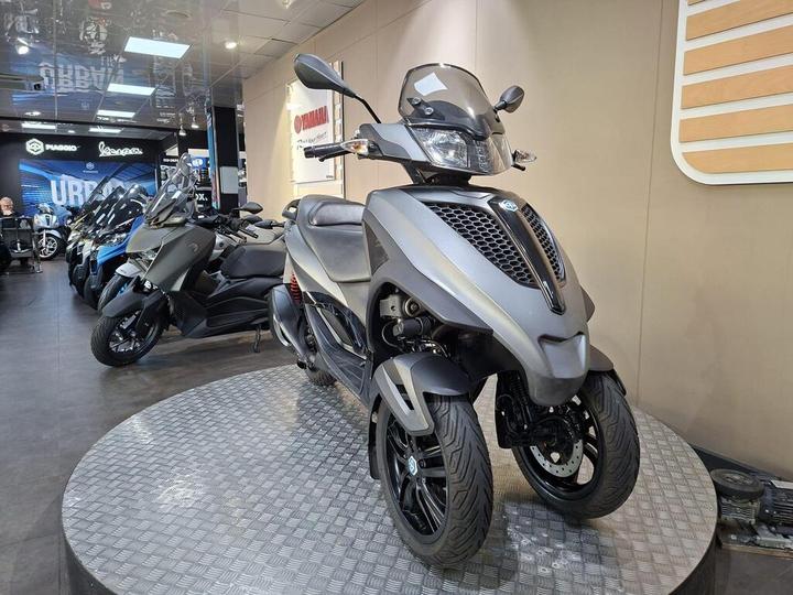 A grey three-wheeled scooter displayed on a circular platform in a motorcycle showroom, with various other scooters visible in the background.