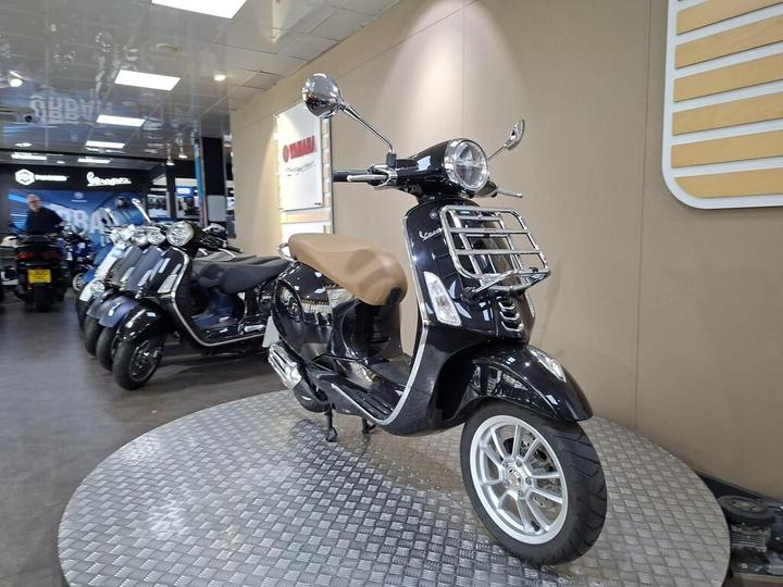 A black Vespa scooter with a brown seat displayed on a circular metal platform in a showroom, with other scooters visible in the background and Yamaha branding on the wall.