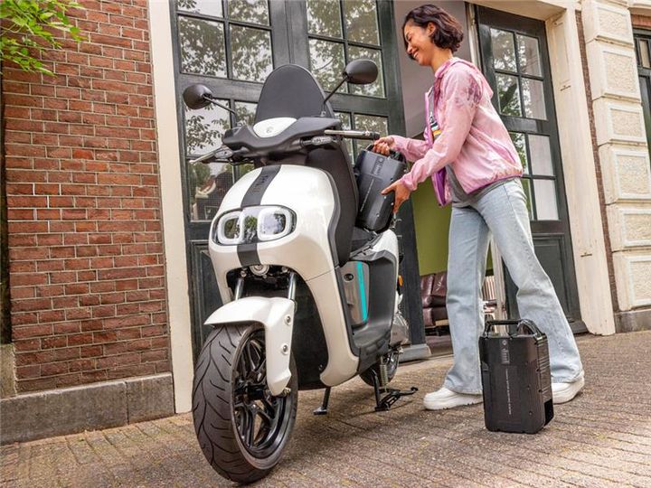 A woman dressed in a light pink jacket and blue jeans is placing a battery pack into an electric scooter parked on a cobblestone street near a brick wall and large doors in the background. A second battery pack sits on the ground next to her.