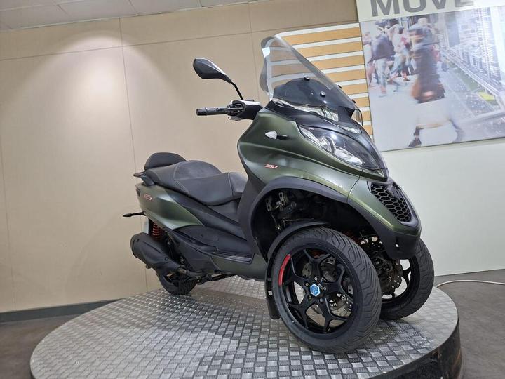 A green three-wheeled scooter displayed on a circular platform, with a transparent windscreen and modern design elements, set against a neutral background.