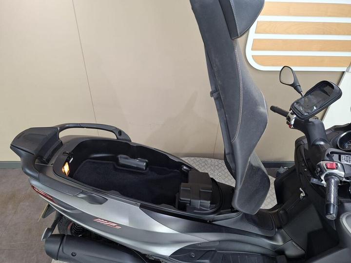 Close-up view of a motorcycle scooter's seat in an upright position, revealing a storage compartment underneath, with a black interior and a dashboard featuring controls and a digital display.