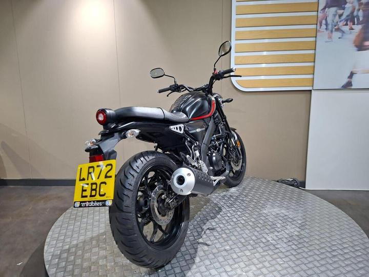 A black motorcycle with red accents is showcased on a round display platform, viewed from the rear-left side. The motorcycle features a prominent exhaust and a visible license plate reading "LR72 EBC." The background includes a muted wall with a simple graphic design.