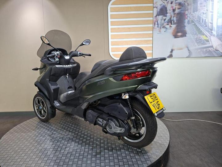 A green three-wheeled scooter displayed on a circular platform, angled to show the rear and side, with a blurred urban scene in the background. The license plate is visible, reading "LS18 YUF."