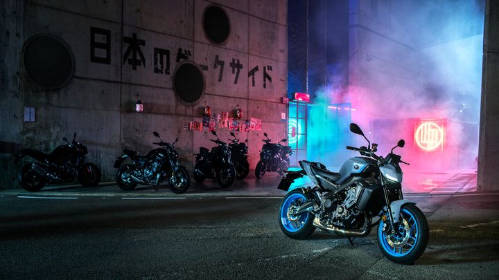 A sleek black motorcycle with blue wheels stands prominently on a street lit by neon lights and surrounded by other motorcycles. The backdrop features a concrete wall with Japanese text and colorful smoke adds to the atmospheric night scene.