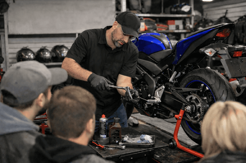 A mechanic demonstrates motorcycle maintenance, using a wrench on a blue bike. Three individuals watch attentively from the foreground, surrounded by tools and bike parts in a workshop setting.