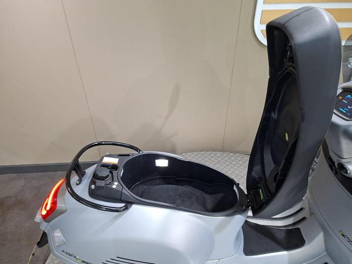 A close-up view of an open storage compartment on a silver scooter, showcasing its interior and the surrounding controls.