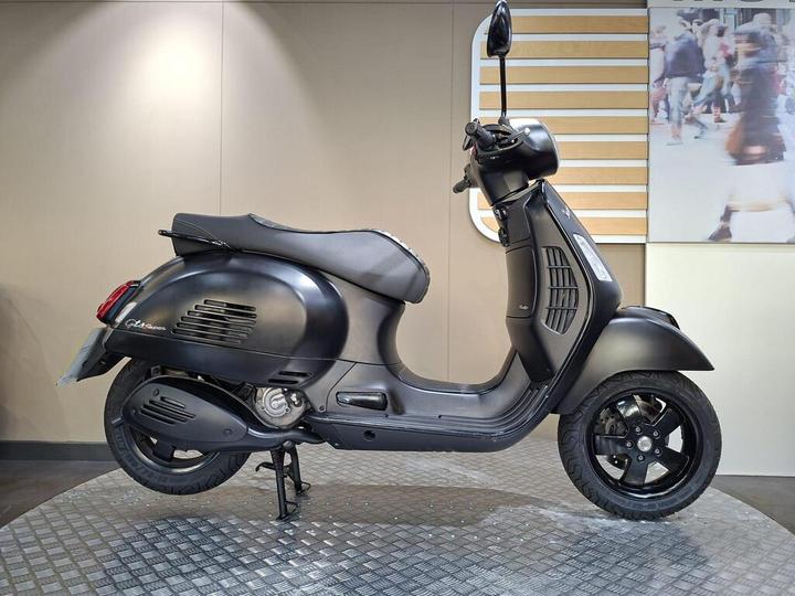 Vespa GTS 300 Super Notte (2019) A stylish black scooter displayed on a diamond-patterned platform against a neutral wall, featuring a sleek design with a rounded front and a low seat.