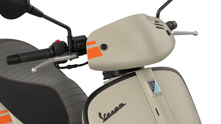 Close-up of a Vespa scooter's handlebars, featuring a beige color scheme, throttle grip, control switches, and a distinctive logo. The seat has a textured design with dark gray and orange accents.