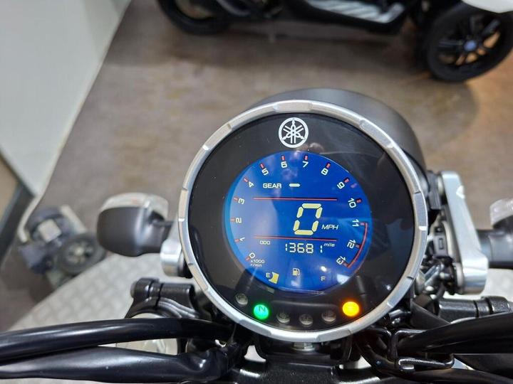 Close-up of a motorcycle dashboard displaying a blue digital speedometer with a speed of 0 MPH, gear indication, and an odometer reading of 1368 miles. The motorcycle's handlebars and controls are visible in the foreground.
