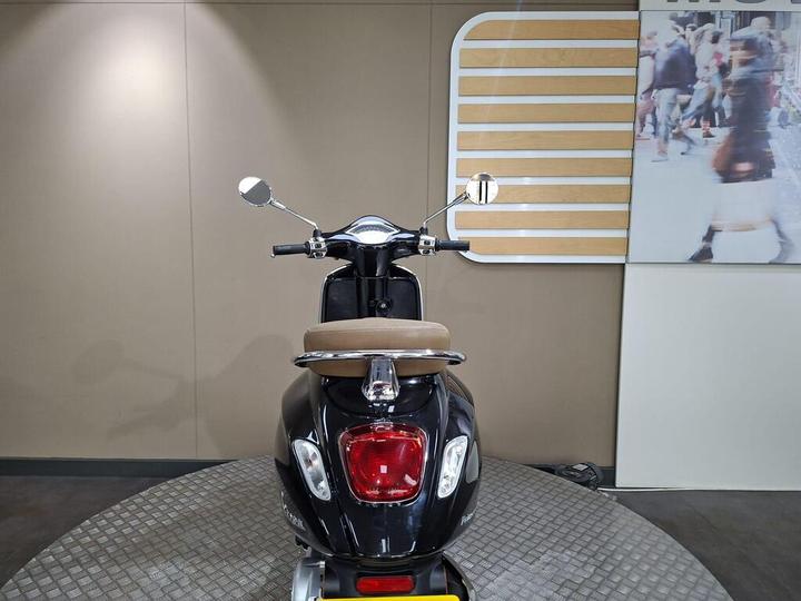A black scooter is displayed from the rear, showcasing its seat and chrome details, against a simple backdrop with a blurred image of pedestrians.