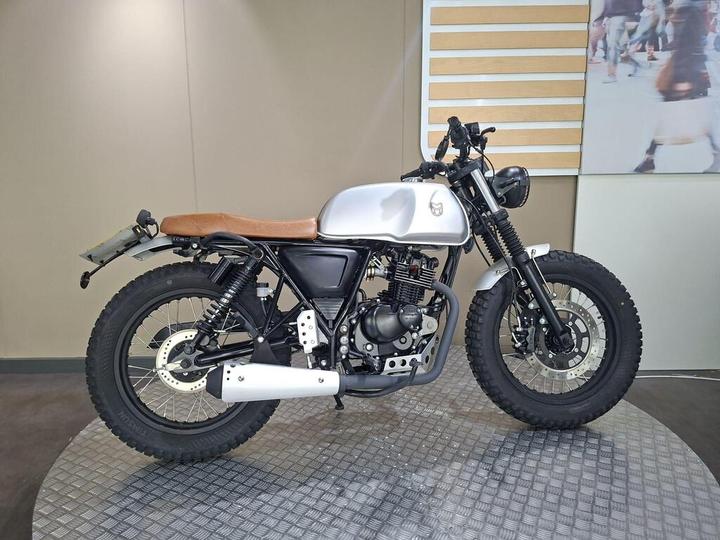 A custom motorcycle with a sleek silver tank, brown leather seat, and rugged tires, positioned on a textured display stand against a neutral background.