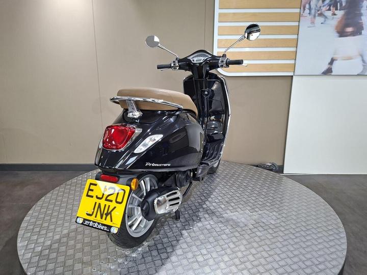 A black scooter with a brown seat is displayed on a circular metal platform, showcasing its rear and side view. The license plate is visible, and the background features a neutral-colored wall.