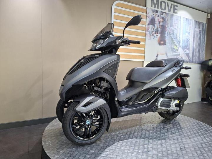 A sleek gray scooter displayed on a circular metal platform in a showroom, featuring two front wheels, a comfortable seat, and modern design elements. The background includes a promotional wall with a cityscape image and the word "MOVE."