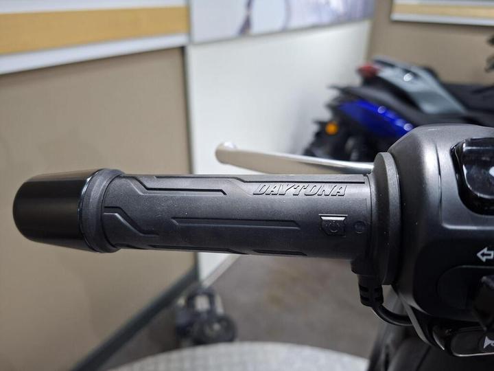 Close-up of a motorcycle throttle grip featuring the word "DAYTONA" embossed on the side, with a blurred background of a motorcycle showroom.