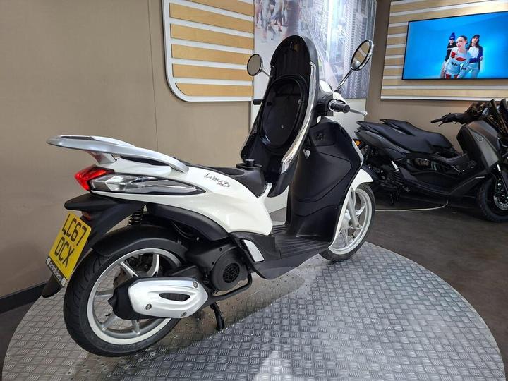 A white and black scooter, the Piaggio Liberty 125, displayed on a textured circular platform, with a blurred background featuring other scooters and a promotional screen.
