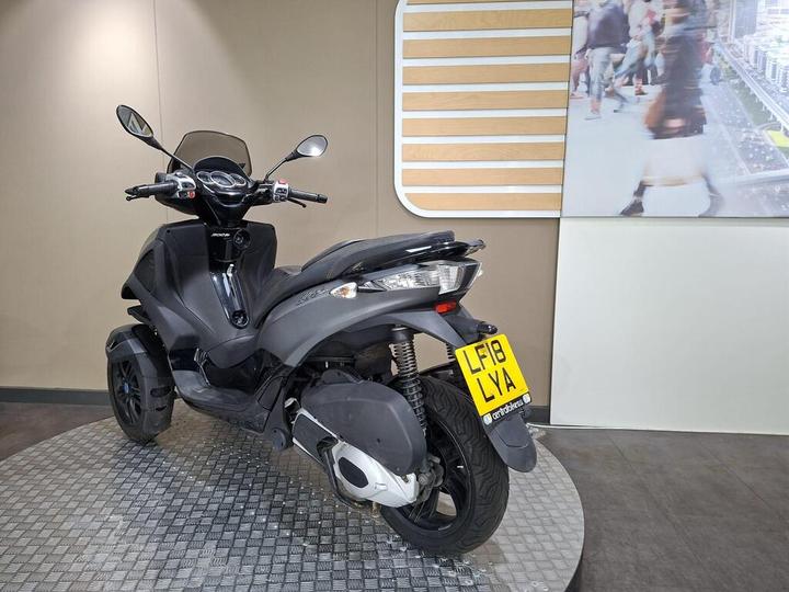 A black three-wheeled scooter parked on a circular platform, with a modern interior background featuring a partially visible digital display of pedestrians. The scooter's license plate reads "LF18 LYA."