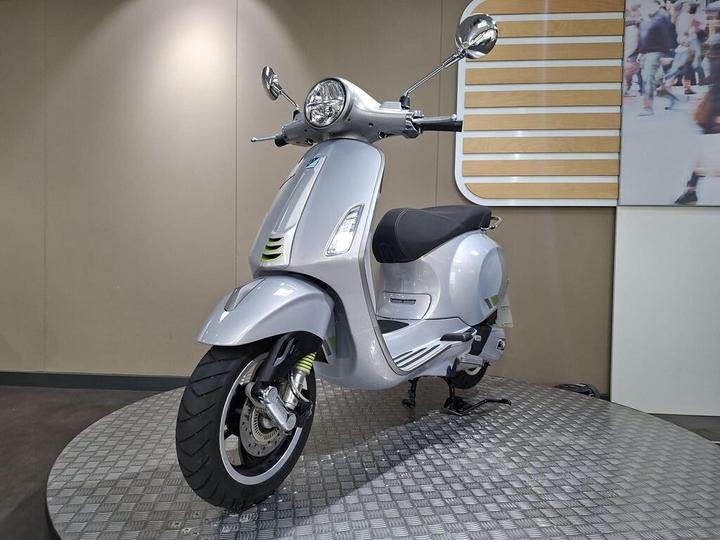 A silver scooter positioned at a slight angle on a textured platform, with a modern showroom backdrop featuring wooden paneling and a blurred crowd image.