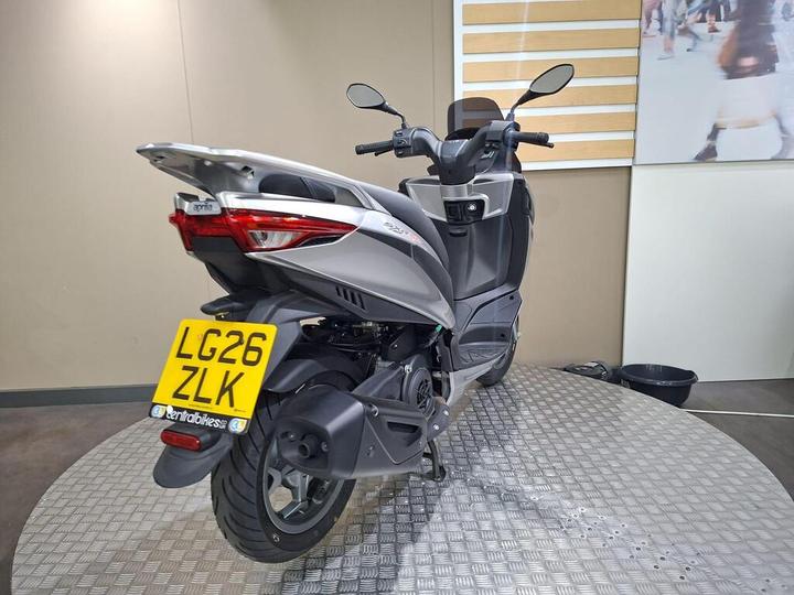 A silver Aprilia scooter positioned on a circular display platform with a textured surface, showcasing its rear view, including the license plate "LG26 ZLK." The background features a plain wall with horizontal stripes.