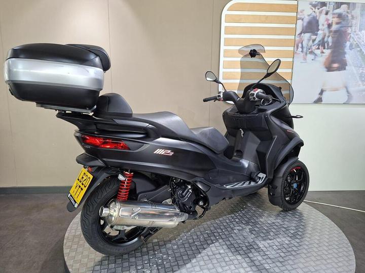 A black three-wheeled scooter with a top case, showcasing a sleek design and red shock absorbers, displayed on a circular platform in a neutral background.