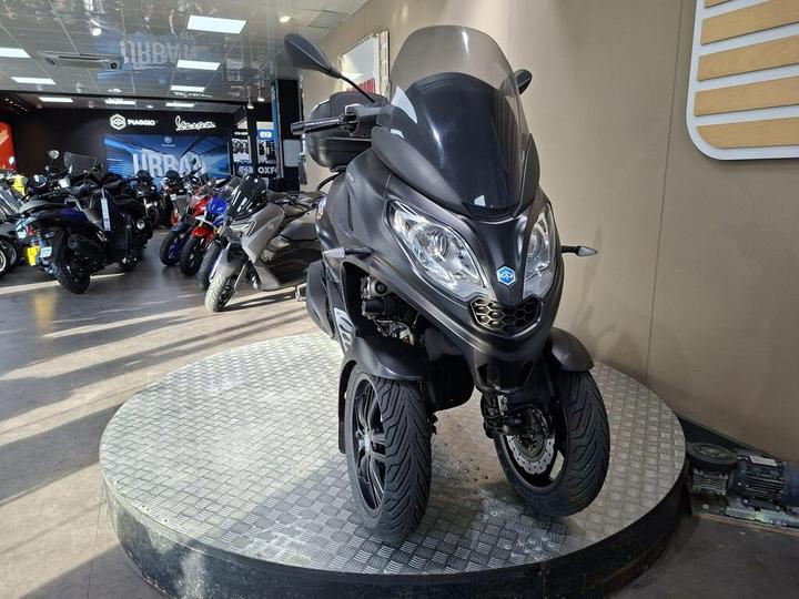 A black three-wheeled scooter displayed on a circular platform in a motorcycle showroom, surrounded by various motorcycles in the background.