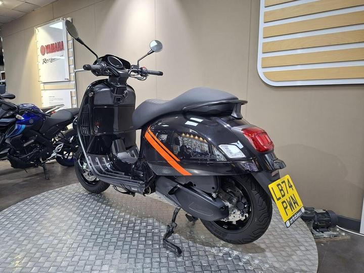 A sleek black scooter with orange accents stands on a textured floor in a Yamaha dealership, with other motorcycles visible in the background.