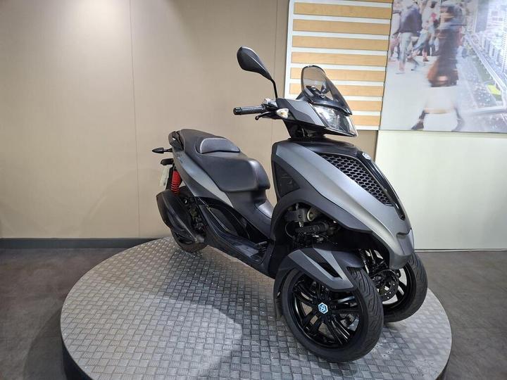 A sleek, modern three-wheeled scooter with a matte gray finish, showcased on a circular display platform against a light-colored wall.