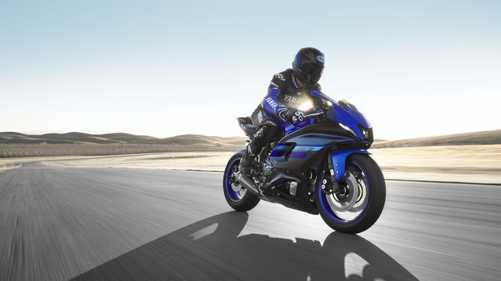A motorcyclist in a black and blue racing suit rides a blue Yamaha R7 on a smooth, winding road with a blurred landscape of rolling hills in the background.
