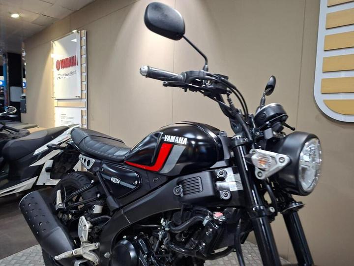 A close-up view of a Yamaha motorcycle, showcasing its black body with red and white accents, featuring a round headlight and a sleek seat, set against a neutral background in a showroom.