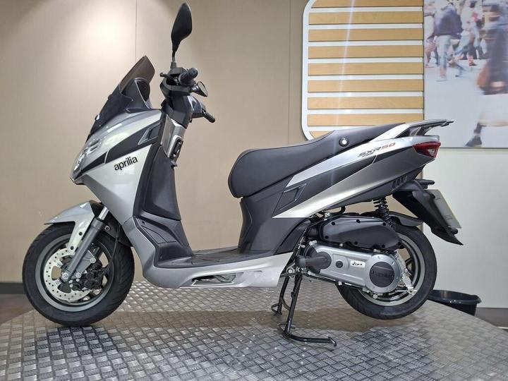 A silver Aprilia SR 50 scooter positioned on a textured floor with a minimalist background featuring a light-colored wall and a graphic design element.