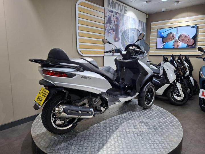 A silver scooter displayed in a showroom, positioned on a circular platform, with various other scooters in the background and a large screen showing smiling people on the wall.