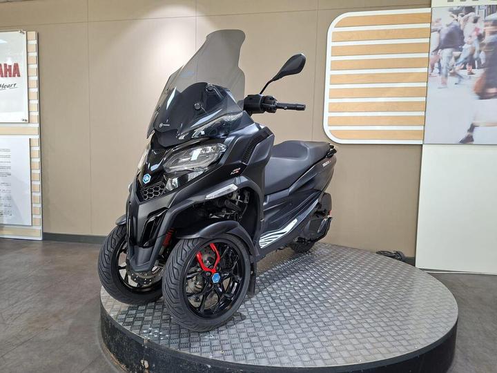 A black three-wheeled scooter with a large windshield is displayed on a round metallic platform in a showroom, with a light-colored wall in the background.