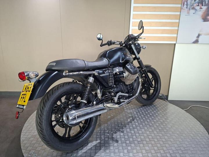 A black Moto Guzzi motorcycle displayed on a round platform, seen from a rear-side angle, featuring chrome exhaust and sleek design elements.