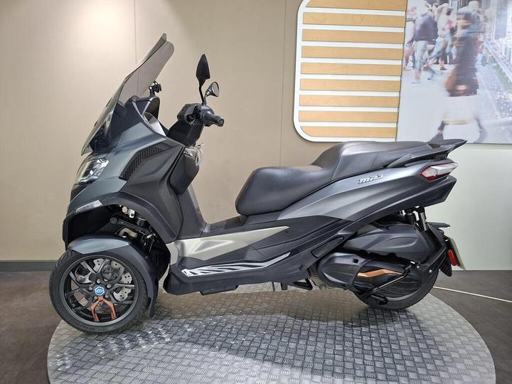 A sleek, gray three-wheeled scooter displayed in a showroom, featuring a protective windshield, dual headlights, and a modern design with sporty accents.