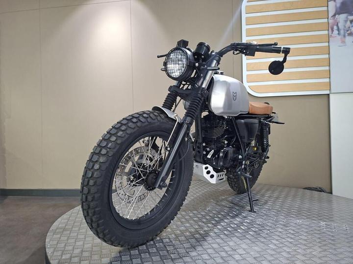 A modern motorcycle with a silver tank, brown leather seat, and chunky tires, displayed on a textured platform in a showroom setting.