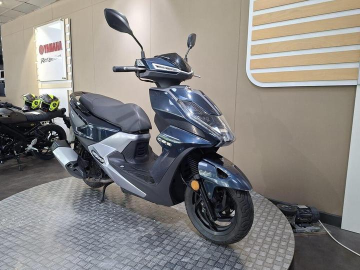 A dark blue scooter is displayed on a textured surface in a showroom, with a Yamaha sign visible in the background. A black motorcycle with green accents is partially seen behind the scooter.