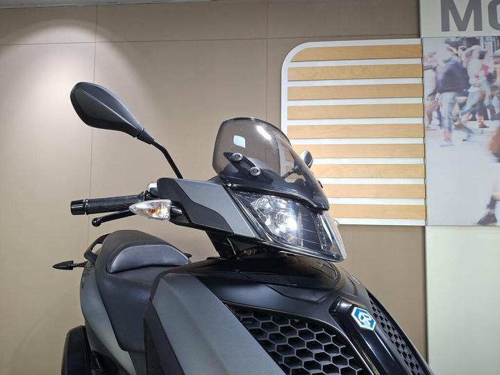 Close-up view of a black scooter with a windscreen and side mirror, displaying sleek design features, set against a neutral background.