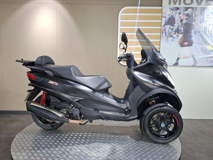 Piaggio MP3 500 Sport (2020) A sleek black three-wheeled scooter displayed on a textured stand, featuring a sport-style design, prominent red suspension springs, and modern detailing, with a light background showcasing a blurred city scene.
