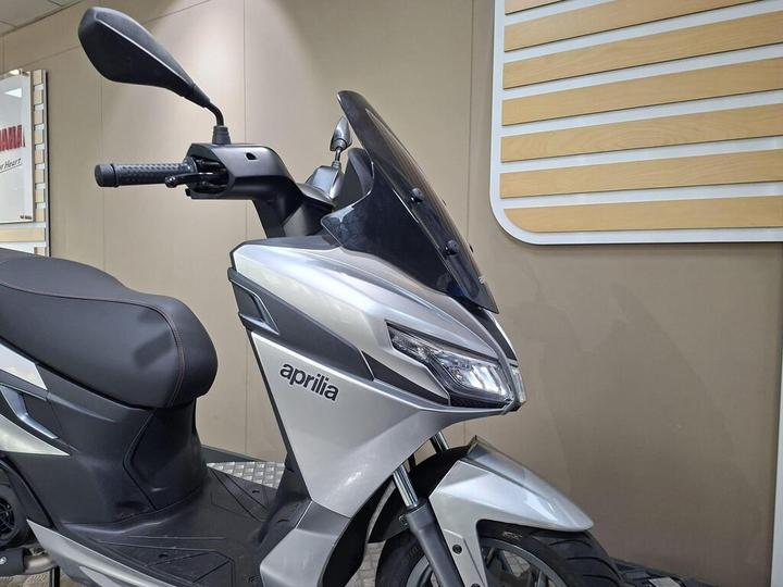 A close-up view of a silver Aprilia scooter, showcasing its sleek design, handlebar, and windshield, with a blurred background of a display wall.