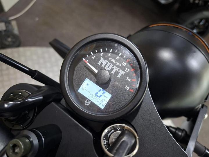 A close-up view of a motorcycle's dashboard featuring a round tachometer labeled "MUTT," displaying RPM readings, and a digital screen showing indicators. The dashboard is attached to a sleek, black motorcycle chassis.