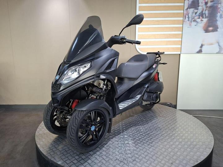 A sleek black three-wheeled scooter displayed on a circular platform, featuring a tinted windshield, comfortable seating, and bold alloy wheels. The background is a soft beige with a subtle pattern, enhancing the scooter's design.
