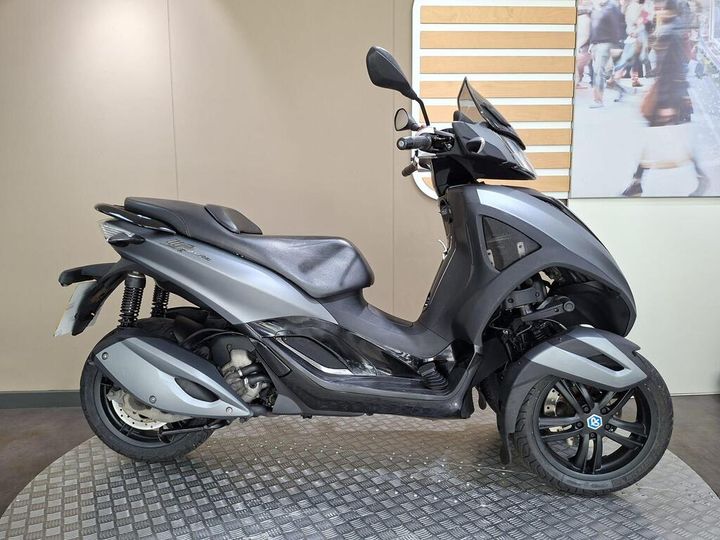 Piaggio MP3 Yourban Sport (2015) A three-wheeled scooter in a sleek silver and black design is displayed on a textured surface against a neutral wall. The scooter features a comfortable seat and modern styling, highlighted by its front and rear wheels.