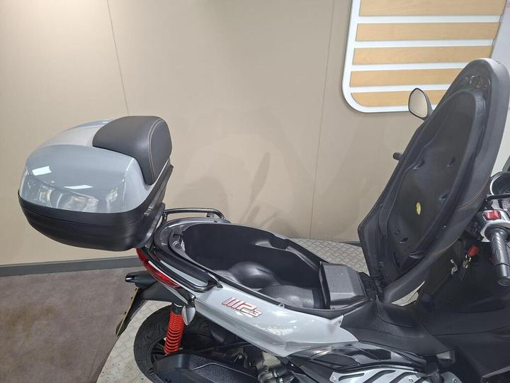 A close-up of a scooter showing an open seat compartment and a gray top case. The scooter features red shock absorbers and a sleek design, set against a neutral background.