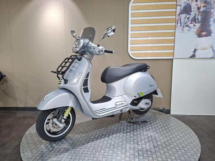 A sleek silver scooter with a black seat and chrome accents, displayed on a circular metallic platform against a neutral background. The scooter features a windscreen, a front rack, and modern design elements.