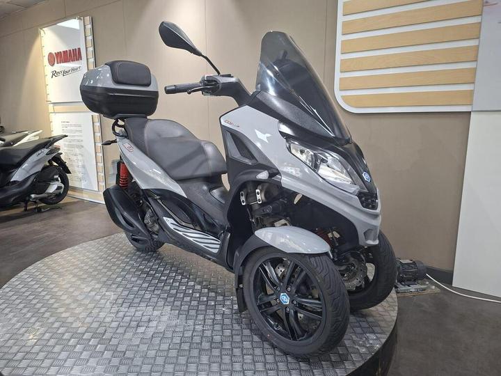 A grey three-wheeled scooter is displayed on a round platform with a textured surface. It features a large windscreen, comfortable seating, and a top case. The background includes a Yamaha branding element and another scooter.