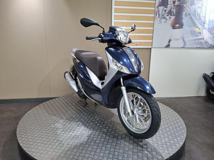 A blue scooter displayed on a rotating pedestal in a showroom, with a beige wall and a cityscape mural in the background.