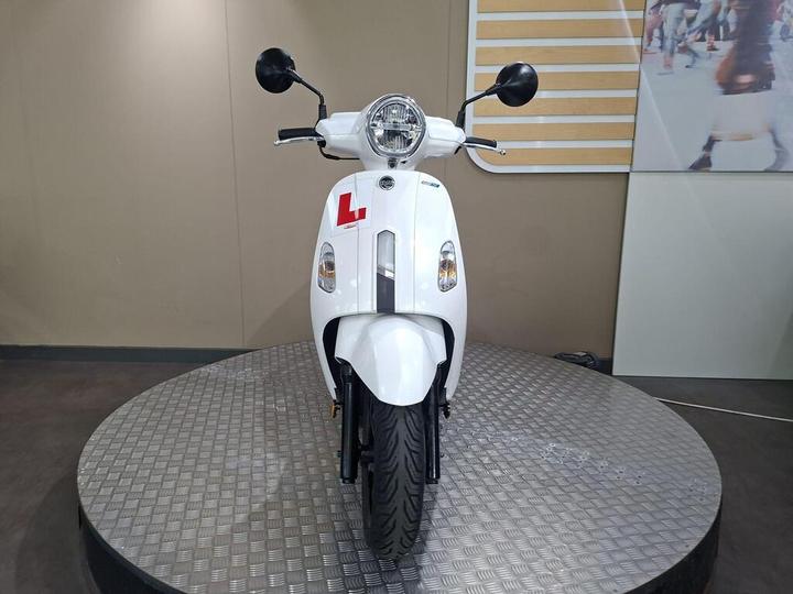 A white scooter with a visible "L" learner's plate is displayed on a circular platform, featuring a round headlight and two mirrors. The background is minimalistic with muted colors.