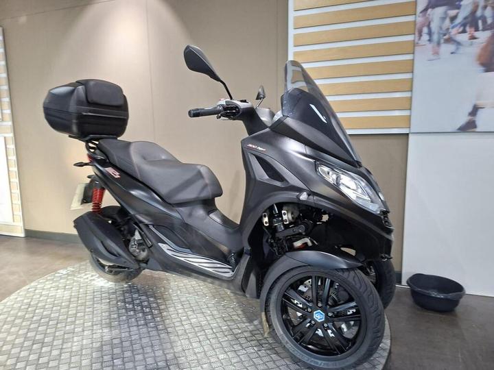 A black three-wheeled scooter is displayed in a modern showroom with a circular patterned floor. It features a large trunk on the back and sleek design elements.