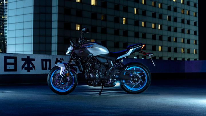 A sleek, modern motorcycle with a silver and black design, illuminated by blue light, parked on a rooftop with a building backdrop and Japanese characters visible on a wall.